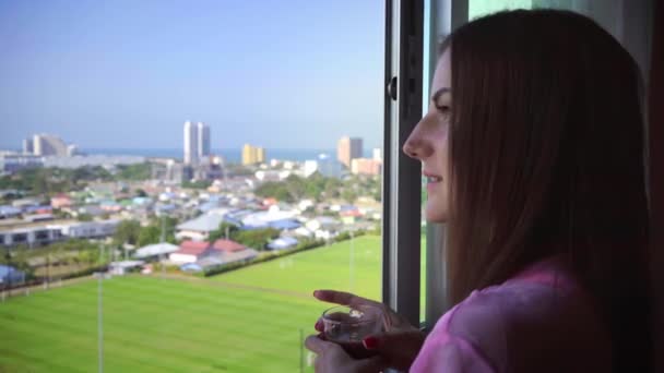 Vrouw in een roze transparante badjas staat en kijkt uit naar het panoramisch venster en drinkt koffie. 4 k Hua Hin, Thailand. — Stockvideo