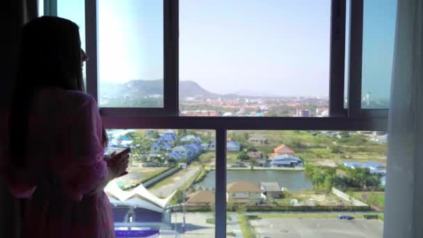 Mujer en una bata transparente rosa se levanta y mira hacia la ventana panorámica y bebe café. 4k Hua Hin, Tailandia . — Vídeos de Stock