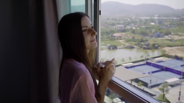 Mujer en una bata transparente rosa se levanta y mira hacia la ventana panorámica y bebe café. 4k Hua Hin, Tailandia . — Vídeos de Stock