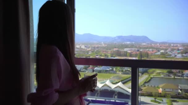 Vrouw in een roze transparante badjas staat en kijkt uit naar het panoramisch venster en drinkt koffie. 4 k Hua Hin, Thailand. — Stockvideo