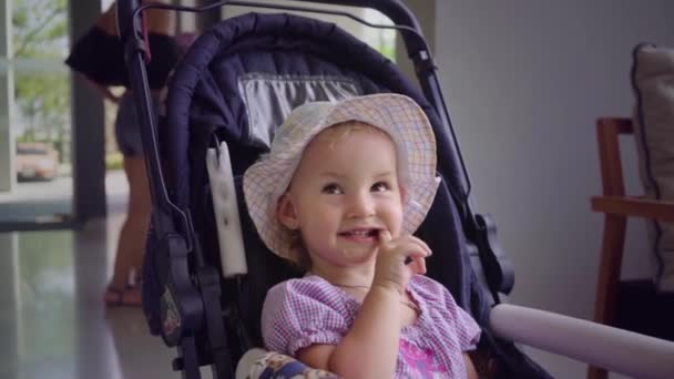 Malá holčička v klobouku sedí v modrém kočárku a olizuje ústa rukou, pomalý pohyb. — Stock video