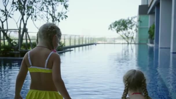 Due sorelle bambine si siedono vicino alla piscina panoramica, vista posteriore, rallentatore — Video Stock