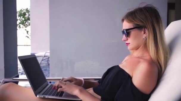 Freelancer kvinna sitter på en solstol nära poolen och arbetar på en bärbar dator, långsam rörelse. — Stockvideo