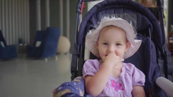 Little girl in a hat sits in a blue stroller and licks her mouth with her hand, slow motion. — Stock Video
