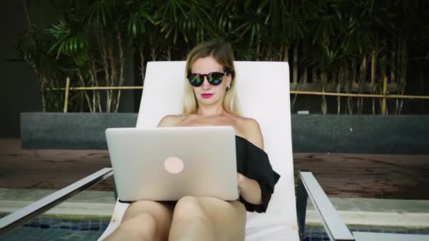 Freelancer woman sitting on a lounger near the pool and working on a laptop, slow motion. — Stock Video