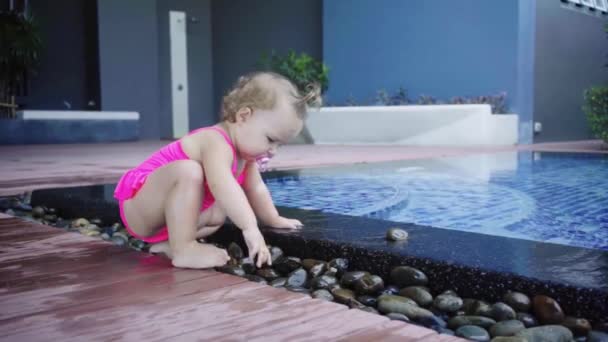 Meisje in een roze badpak zit in het ondiepe water van het zwembad. Het kind gooit stenen vanaf het zwembad, slow-motion. — Stockvideo