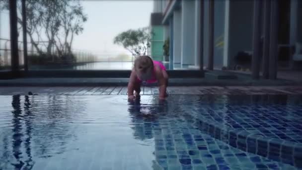 Meisje in een roze badpak zit in het ondiepe water van het zwembad. Het kind gooit stenen vanaf het zwembad, slow-motion. — Stockvideo