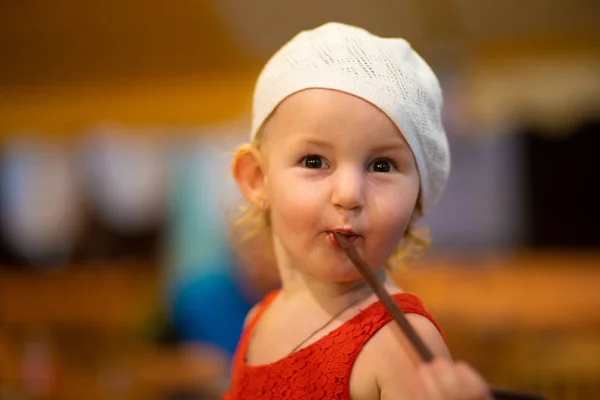 Portré egy gyermek lány egy fehér kalapot, és a piros ruha, a gyermek három éves iszik a szalma. — Stock Fotó