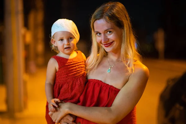 Mother holds daughter in her arms, mother with daughter is dressed in red dresses, child in white beret, at night. — Stock Photo, Image