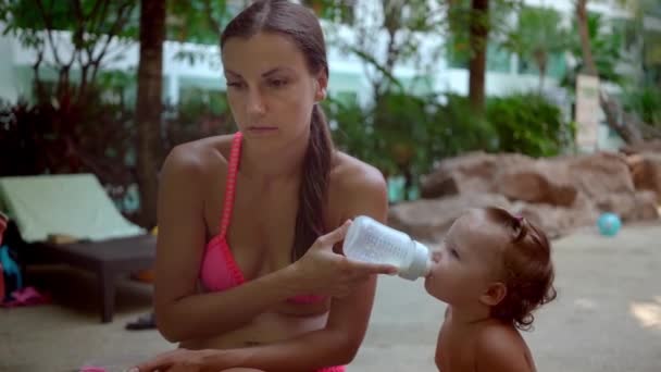 Moe en gemarteld moeder feeds baby meisje dochter met een fles melk in een tropische tuin, langzame motie. — Stockvideo