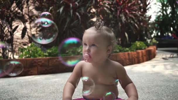 Portrait baby age one year old, girl sits with pacifier and looks at soap bubbles on the street in the garden, slow motion. — Stock Video