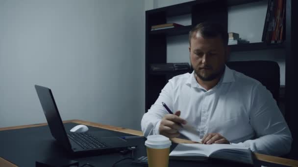 Un uomo d'affari barbuto siede in ufficio a un computer portatile, un uomo conta su una calcolatrice e prende appunti, pianificazione del bilancio, entrate, rallentatore . — Video Stock