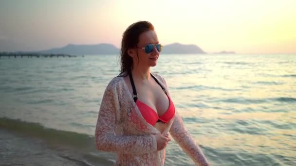 Mulher bonito com sardas em um maiô rosa e vestido branco em óculos de sol em uma praia de areia branca com palmeiras andando ao longo da praia e sorrindo — Vídeo de Stock