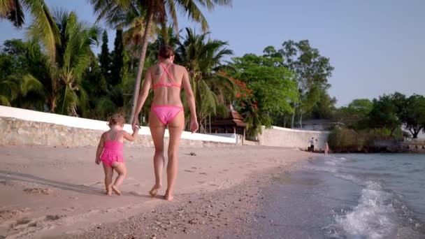 Мама и маленькая дочь в розовых купальниках прогуливаются вдоль берега моря, вид сзади, замедленное движение . — стоковое видео