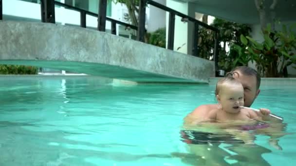 Sevimli küçük çocuk ve babası havuzda yüzme dersi sahip. Babası kızı onun kollarında tutar ve ona sarılarak. Küçük kız mutlu bir şekilde gülümsüyor ve çalış — Stok video