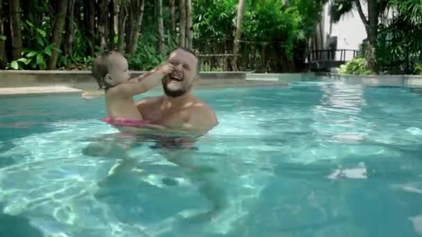 Lindo niño y su padre teniendo una clase de natación en la piscina. El padre sostiene a la hija en sus brazos y la abraza. La niña sonríe alegremente y juega — Vídeos de Stock