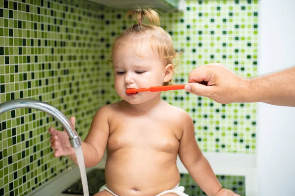 Söta barn girl borsta hennes tänder i badrummet. Söt baby sitter i diskhon. Pappa hjälper till att borsta barnets tänder — Stockfoto