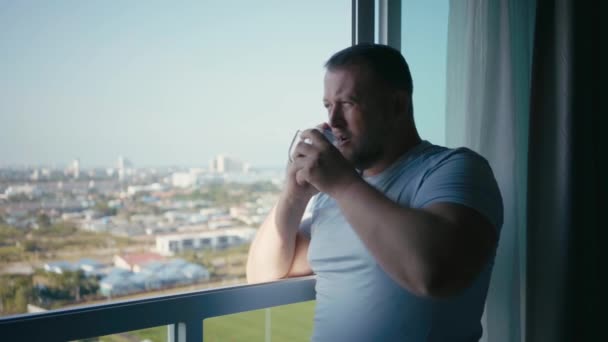 Mężczyzna pije kawy i patrzy przez panoramiczne okna uśmiechy rankiem, zwolnionym tempie — Wideo stockowe