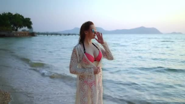 Ładna kobieta z białej sukni w okulary na piaszczystej plaży z palmami, spacery wzdłuż plaży i uśmiechnięte i piegi w różowy strój kąpielowy — Wideo stockowe