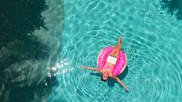 透明なターコイズプールでラップトップを持つインフレータブル大きなドーナツで泳ぐ若いブルネットの女性の航空写真. — ストック写真