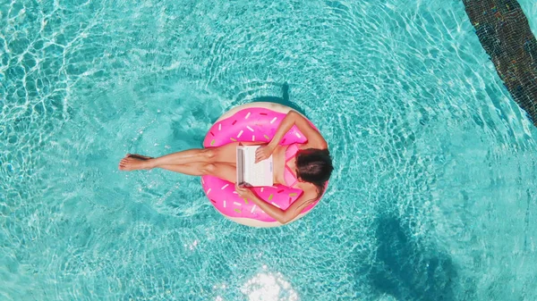 透明なターコイズプールでラップトップを持つインフレータブル大きなドーナツで泳ぐ若いブルネットの女性の航空写真. — ストック写真
