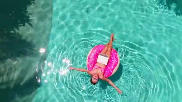 Luchtfoto van een jonge brunette vrouw zwemmen op een opblaasbare grote donut met een laptop in een transparant turquoise zwembad. — Stockvideo