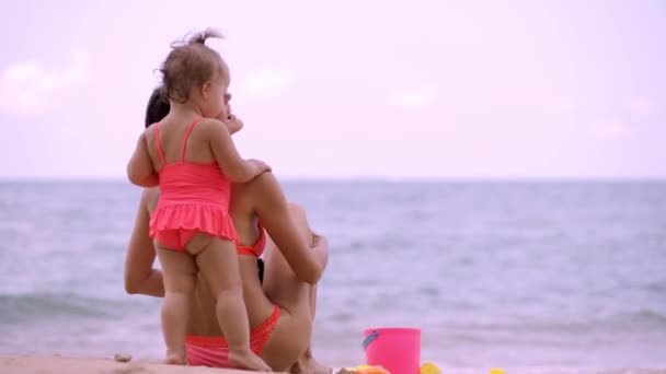 Mutter und Tochter in rosa Badeanzügen am Ufer eines schönen Strandes, der im Sand mit Kinderspielzeug spielt. Mutter und Kind blicken in die Ferne auf das blaue Meer — Stockvideo