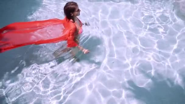 ピンクの長いドレスを着た女性は、水の下の青いプールで泳ぎます。ピンクの生地は、水の中で開発します. — ストック動画