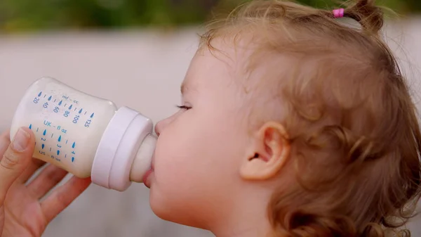 Ένα μικρό όμορφο κορίτσι με μπλε μάτια πίνει γάλα από ένα μπουκάλι. Το παιδί κρατά το μπουκάλι. — Φωτογραφία Αρχείου