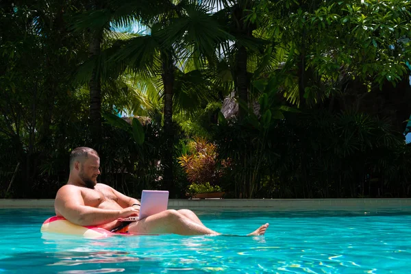 Чоловік з надувним кільцем у воді в басейні працює на комп'ютері. Забавна товста людина в плавальному колі з рожевим ноутбуком. концепція фрілансу — стокове фото