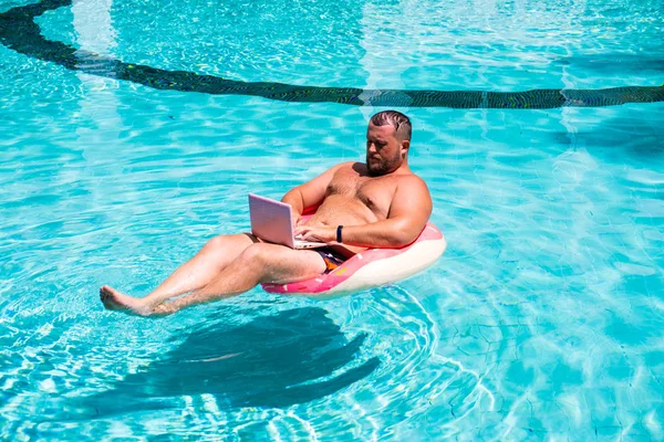 Фрілансер з надувним кільцем у воді в басейні працює на комп'ютері. Забавна товста людина в плавальному колі з рожевим ноутбуком. концепція фрілансу — стокове фото