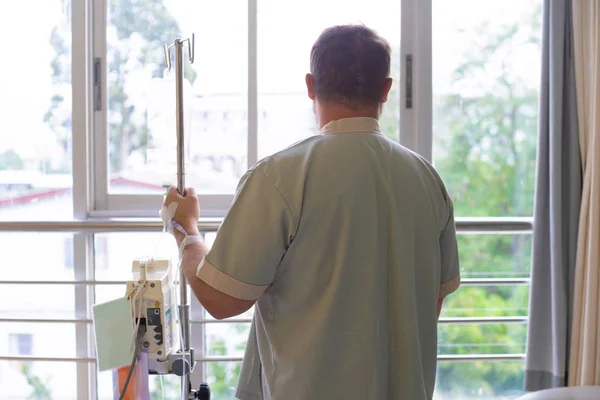 Ασθενής με ένα σταγονόμετρο κοιτάζει έξω από το παράθυρο του νοσοκομείου και χαμόγελα. πίσω όψη. Έννοια της υγειονομικής περίθαλψης. — Φωτογραφία Αρχείου