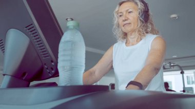 Kıdemli kadın spor salonunda bir koşu bandı üzerinde meşgul. sarışın kadın koşu bandı üzerinde kulaklıklar ile müzik dinler