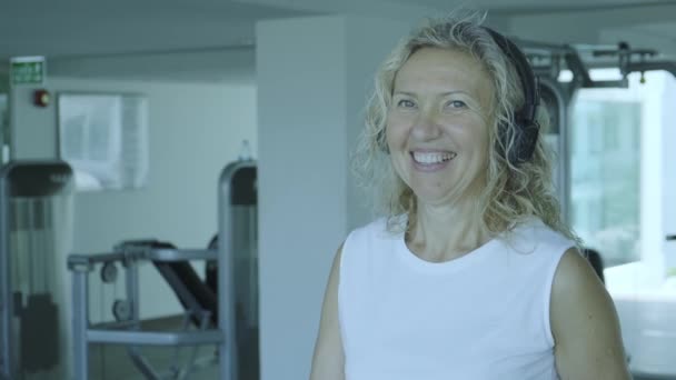 Femme âgée est engagée sur un tapis roulant dans la salle de gym. blonde femme écoute de la musique avec des écouteurs sur le tapis roulant — Video