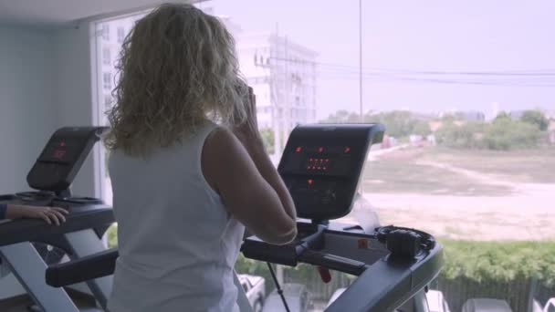 Mujer mayor se dedica a una cinta de correr en el gimnasio. mujer hablando por teléfono haciendo la cinta de correr — Vídeos de Stock