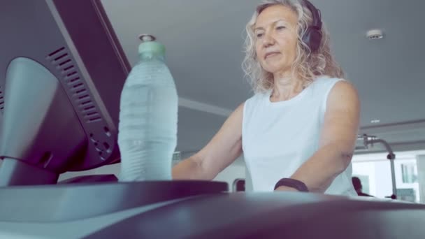 Donna anziana è impegnata su un tapis roulant in palestra. bionda femmina ascolta musica con cuffie sul tapis roulant — Video Stock