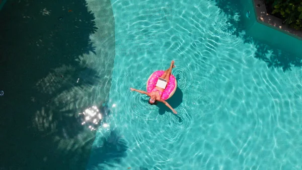 スイミングプール内の水の中にインフレータブルリング上のピンクのノートパソコンと美しい若い女性。フリーランス、ビジネスと旅行のコンセプト。上空表示 — ストック写真