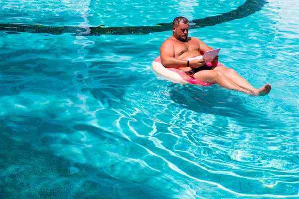 Фрілансер з надувним кільцем у воді в басейні працює на комп'ютері. Забавна товста людина в плавальному колі з рожевим ноутбуком. концепція фрілансу — стокове фото