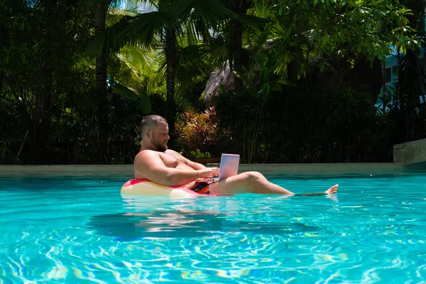 Freelancer met een opblaasbare ring in het water in het zwembad werkt op de computer. Grappige Fat Man in zwemmen cirkel met roze laptop. freelance concept — Stockfoto