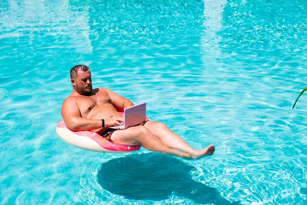 male with an inflatable ring in the water in the pool works on the computer. Funny Fat Man in Swimming Circle with Pink Laptop. freelance concept
