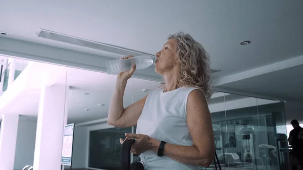 Senior vrouw drinkt water uit drinkwater in de sportschool — Stockfoto