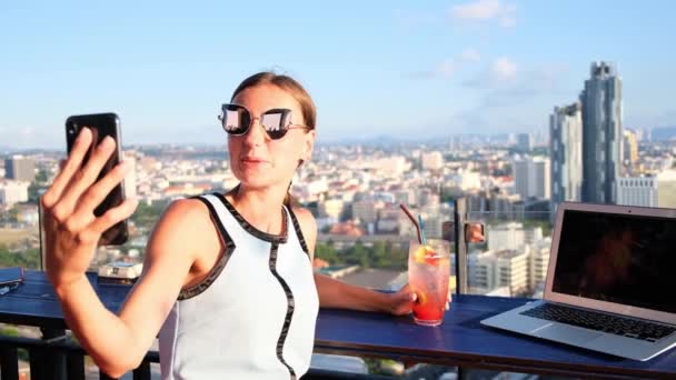 Hembra hace selfie en un café en el techo de un rascacielos. chica sentada con un ordenador portátil y un cóctel naranja en contra de la vista panorámica — Vídeos de Stock
