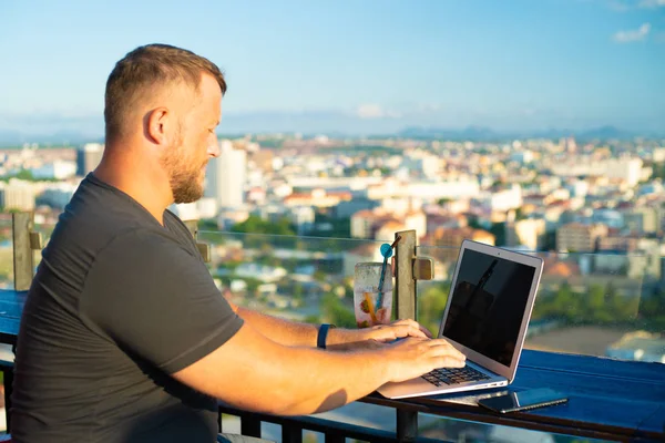 Maschio che lavora su un computer portatile in un caffè sul tetto con una bella vista panoramica. uomo bere un cocktail e lavorare su un computer . — Foto Stock