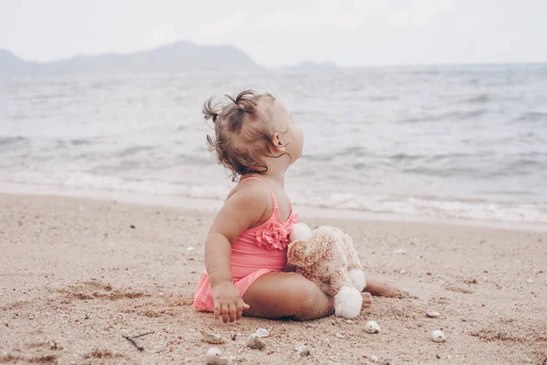 Pensieroso bambina abbracciando orsacchiotto e guardando lontano mentre seduto sulla riva del mare . — Foto Stock