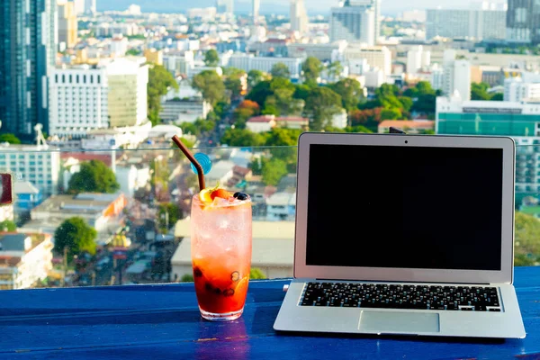 Ноутбук і келих літнього коктейлю на дерев'яному столі, на яскравому красивому панорамному тлі міста . — стокове фото