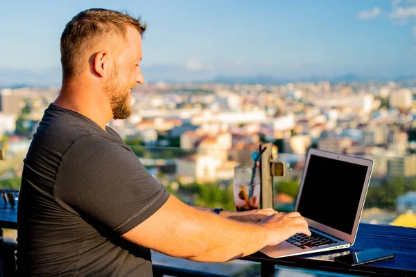 Maschio che lavora su un computer portatile in un caffè sul tetto con una bella vista panoramica. uomo bere un cocktail e lavorare su un computer . — Foto Stock