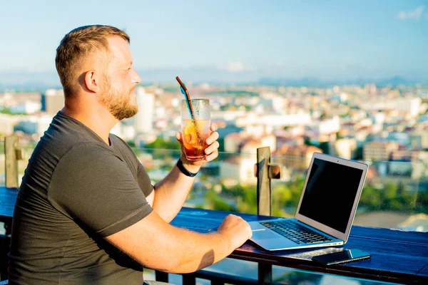 Maschio parlare al telefono e lavorare su un computer portatile in un caffè sul tetto con vista panoramica. uomo bere un cocktail e lavorare su un computer . — Foto Stock