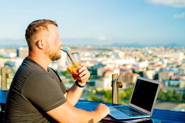 Maschio parlare al telefono e lavorare su un computer portatile in un caffè sul tetto con vista panoramica. uomo bere un cocktail e lavorare su un computer . — Foto Stock