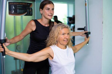 Kıdemli kadın kişisel bir antrenör ile spor salonunda bir simülatör üzerinde meşgul. kız spor salonunda anne yardımcı olur
