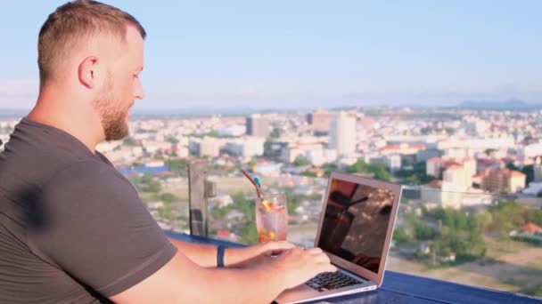 Mann arbeitet an einem Laptop in einem Café auf dem Dach mit schönem Panoramablick. Mann trinkt einen Cocktail und arbeitet am Computer. — Stockvideo